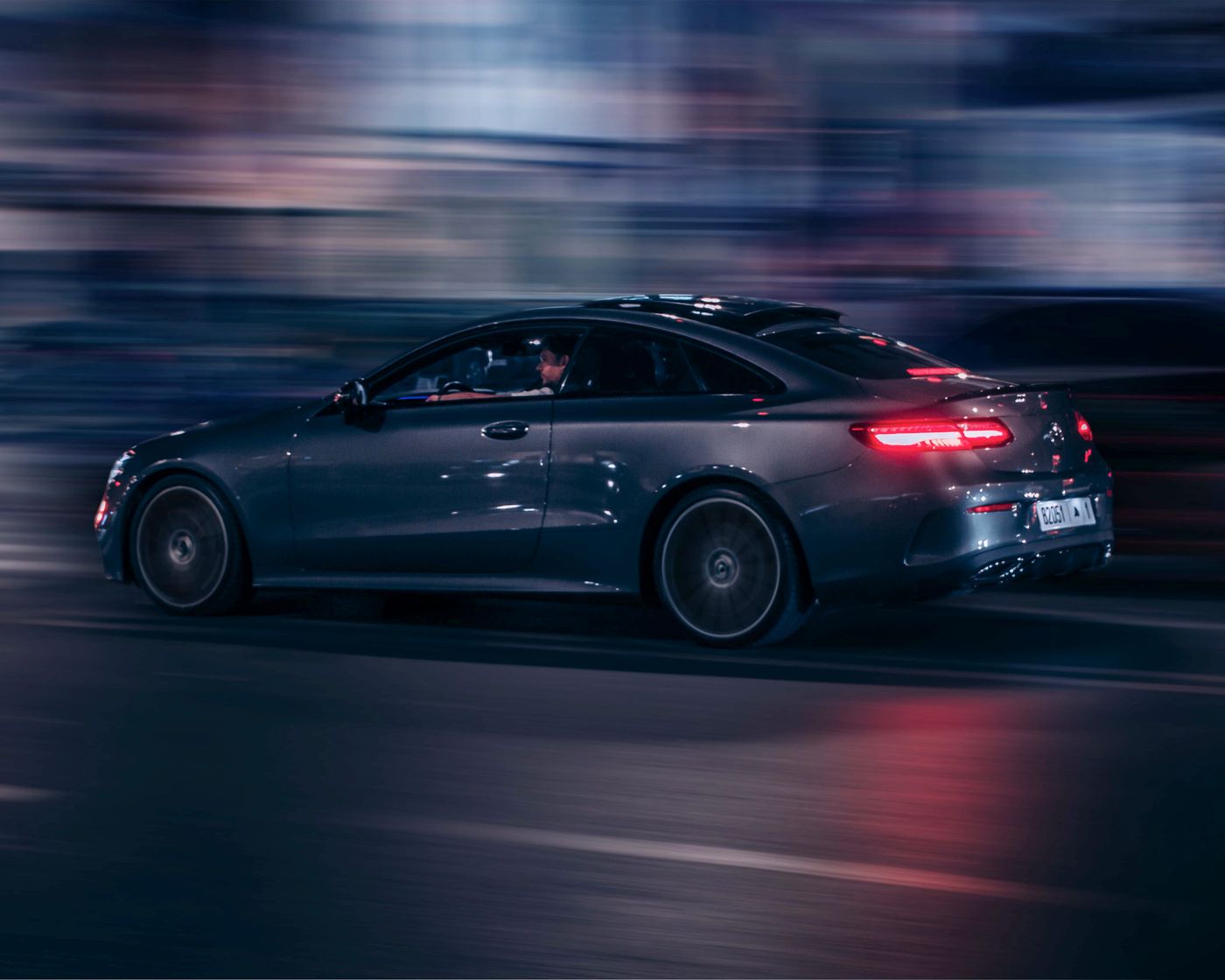 A dark Mercedes saloon moving through a city street at night, shop lights streaked behind it